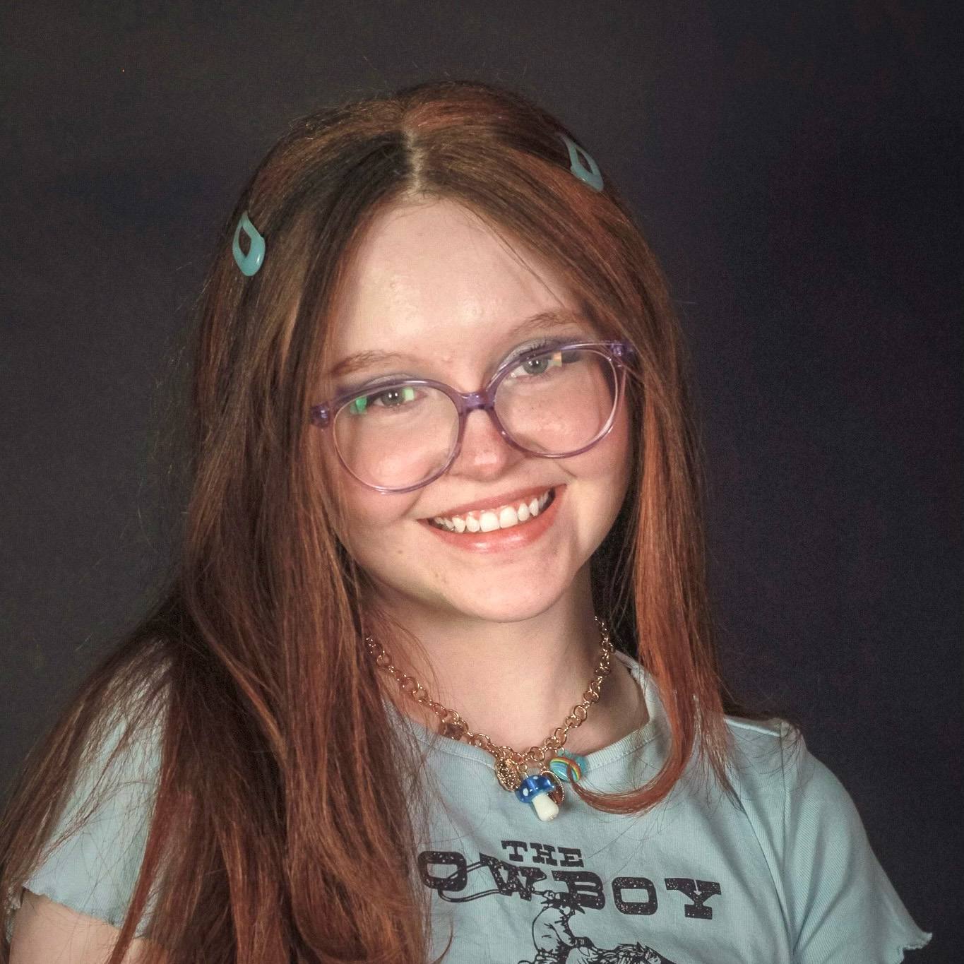 Annika Godwin posing for a headshot with two green clips in her red hair wearing purple rimmed glasses and a light green shirt.