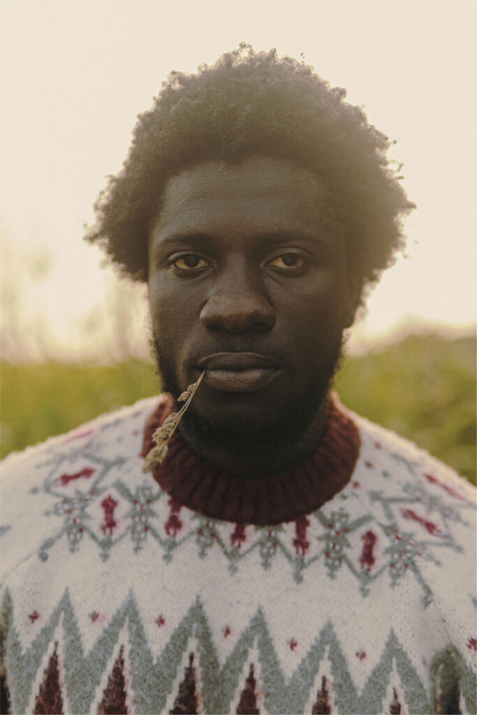 Man wearing a patterned red, white and gray sweater with a twig of hay hanging out of his mouth, standing in a field.