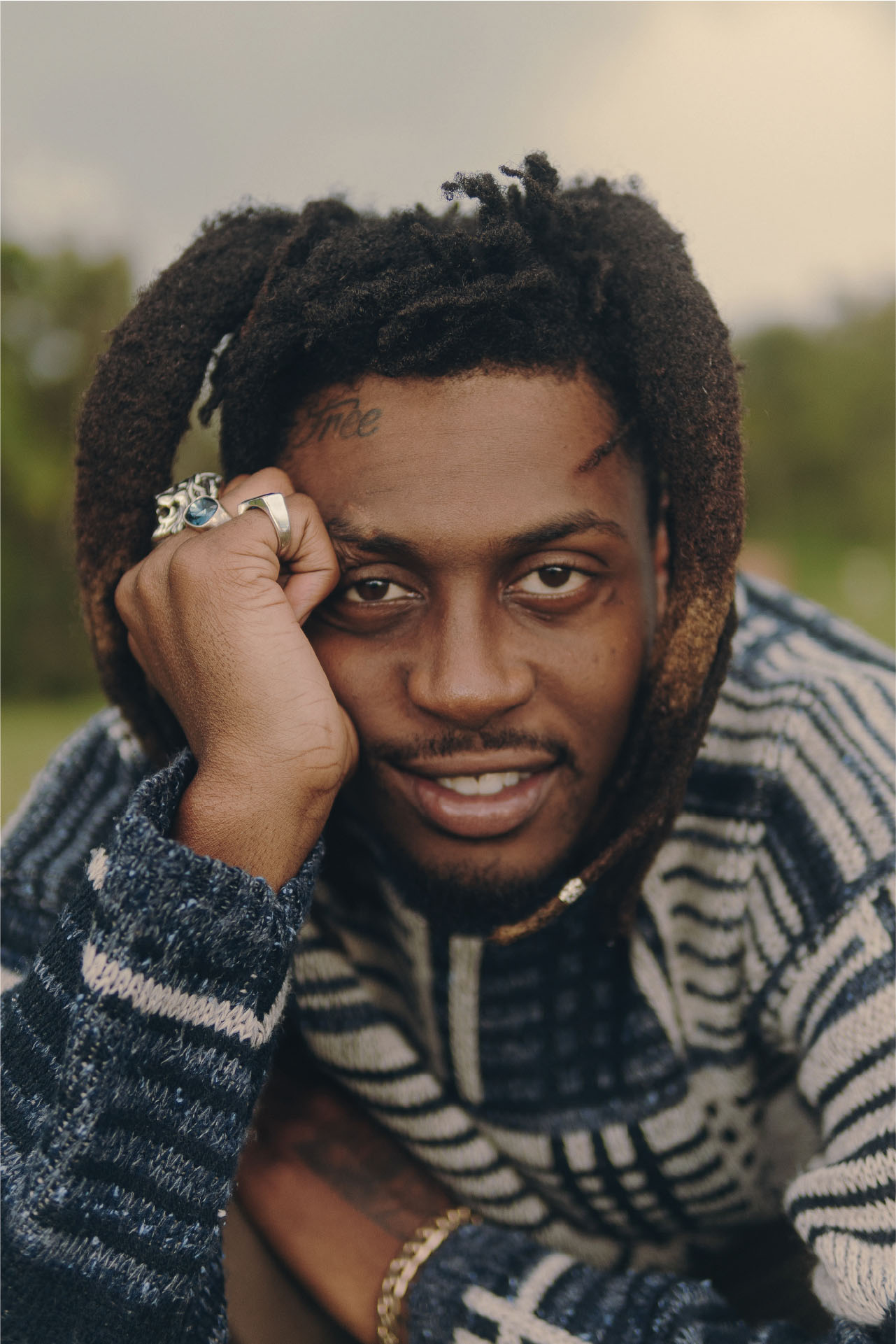 Man posing with a patterned textured blue and white sweater, leaning on his hand with large rings on every finger.