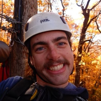 Joeseph Lucey posing for a headshot wearing a white helmet in front of autumn yellow leaves in the trees.