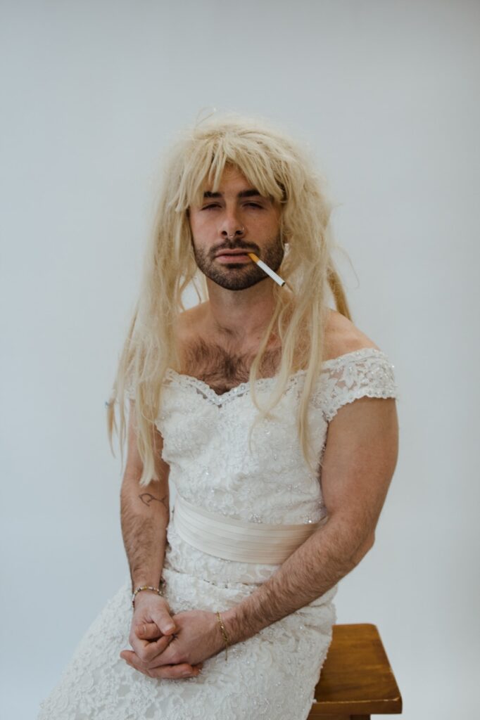 Justin Darnell posing with a blonde wig and cigarette while wearing a white dress by Justin Murphy Justin Darnell posing with a blonde wig and cigarette while wearing a white dress